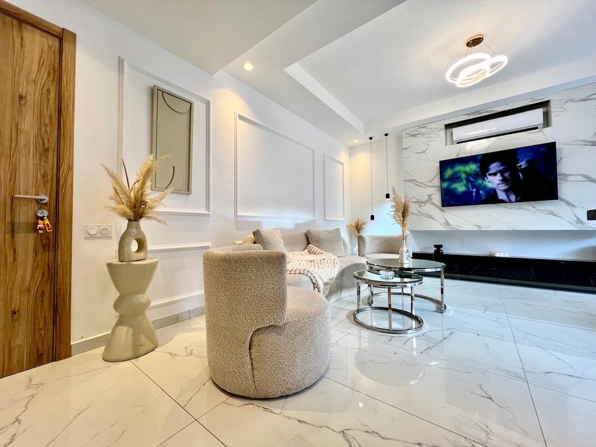 A modern living room is presented with marble flooring and contemporary decor. Plush chairs are arranged around a glass coffee table, while a wall-mounted television adds a focal point. Natural light is filtered through the space, enhancing the minimalist design.