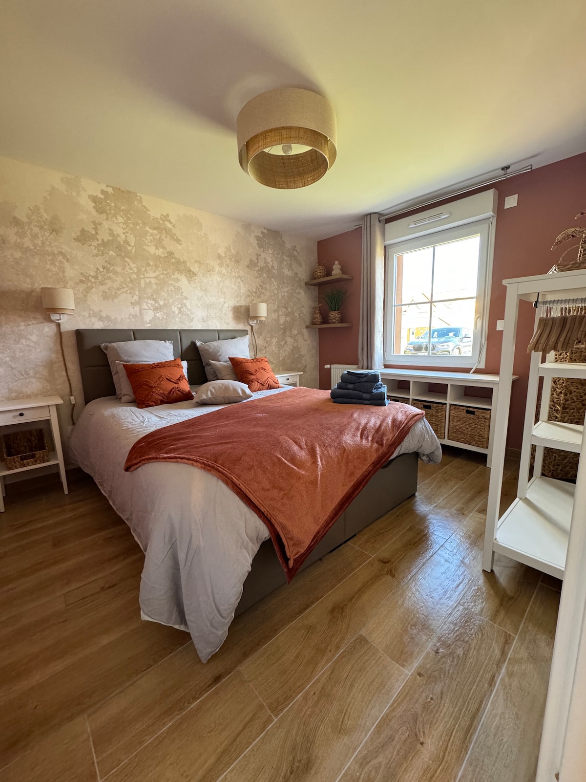 The bedroom features a spacious bed with decorative pillows and a cozy throw draped over the side. Light wood flooring complements the warm-toned walls adorned with subtle patterns. A window allows natural light to brighten the room, while bedside tables and shelving provide additional storage.