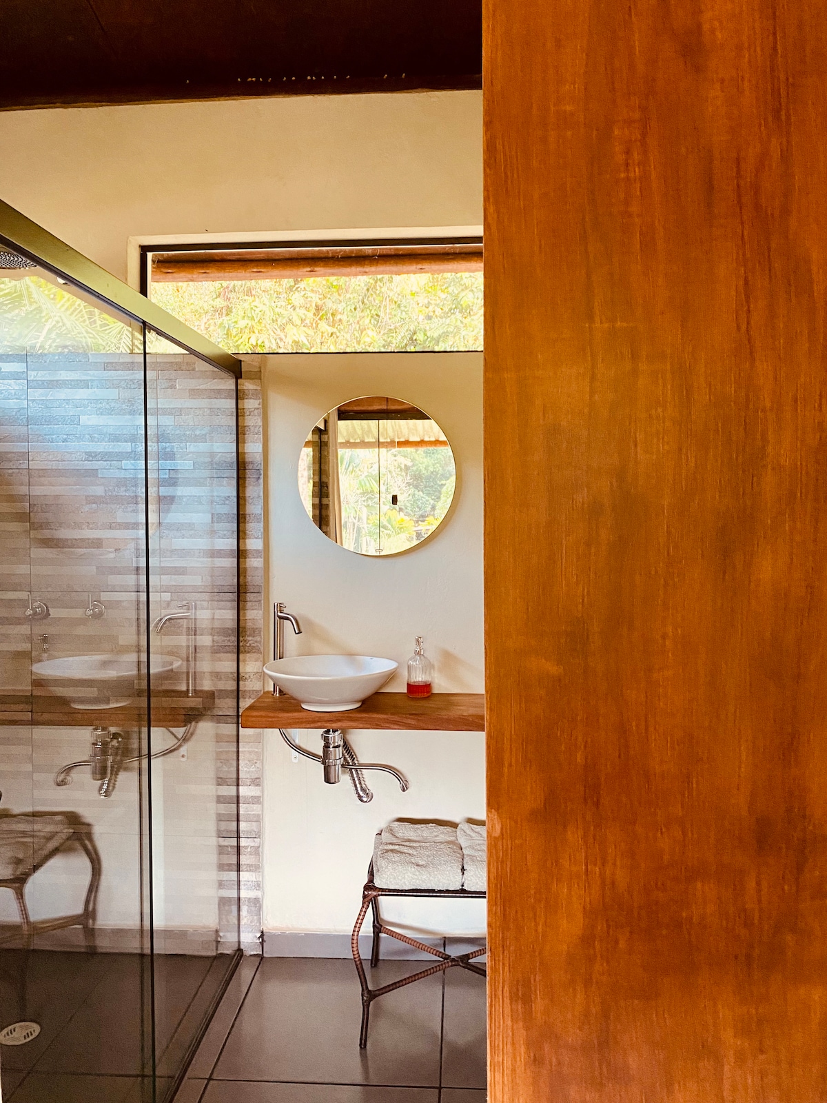 A modern bathroom features a glass shower enclosure and a sleek sink on a wooden counter. Natural light floods the space through a large window, while a round mirror reflects the interior design elements. A small stool sits beside the shower, adding functionality.
