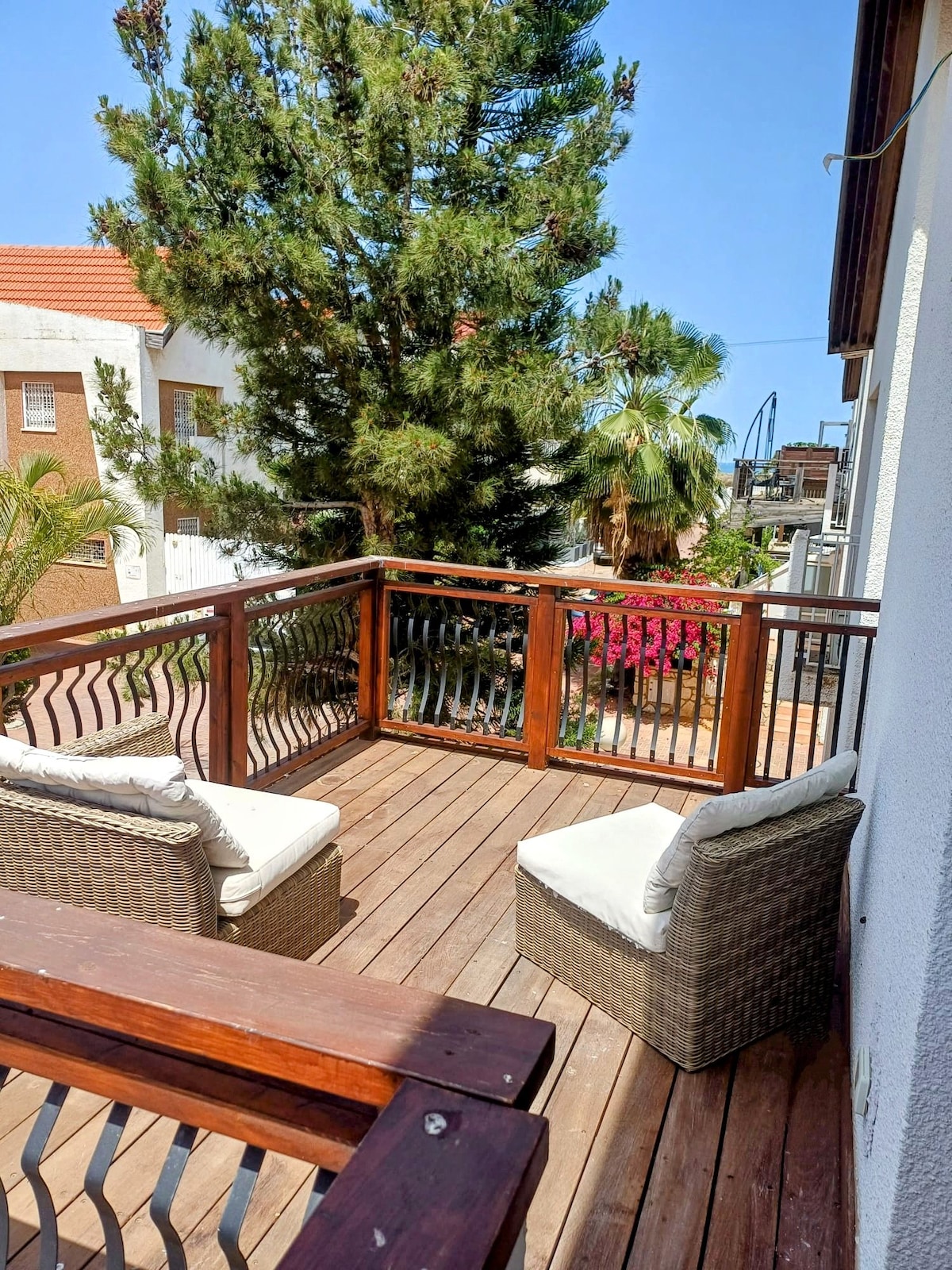 A wooden deck features two cozy wicker chairs, inviting relaxation. Lush greenery and vibrant flowers surround the area, enhancing the outdoor setting. The sky is clear and bright, contributing to a serene atmosphere.
