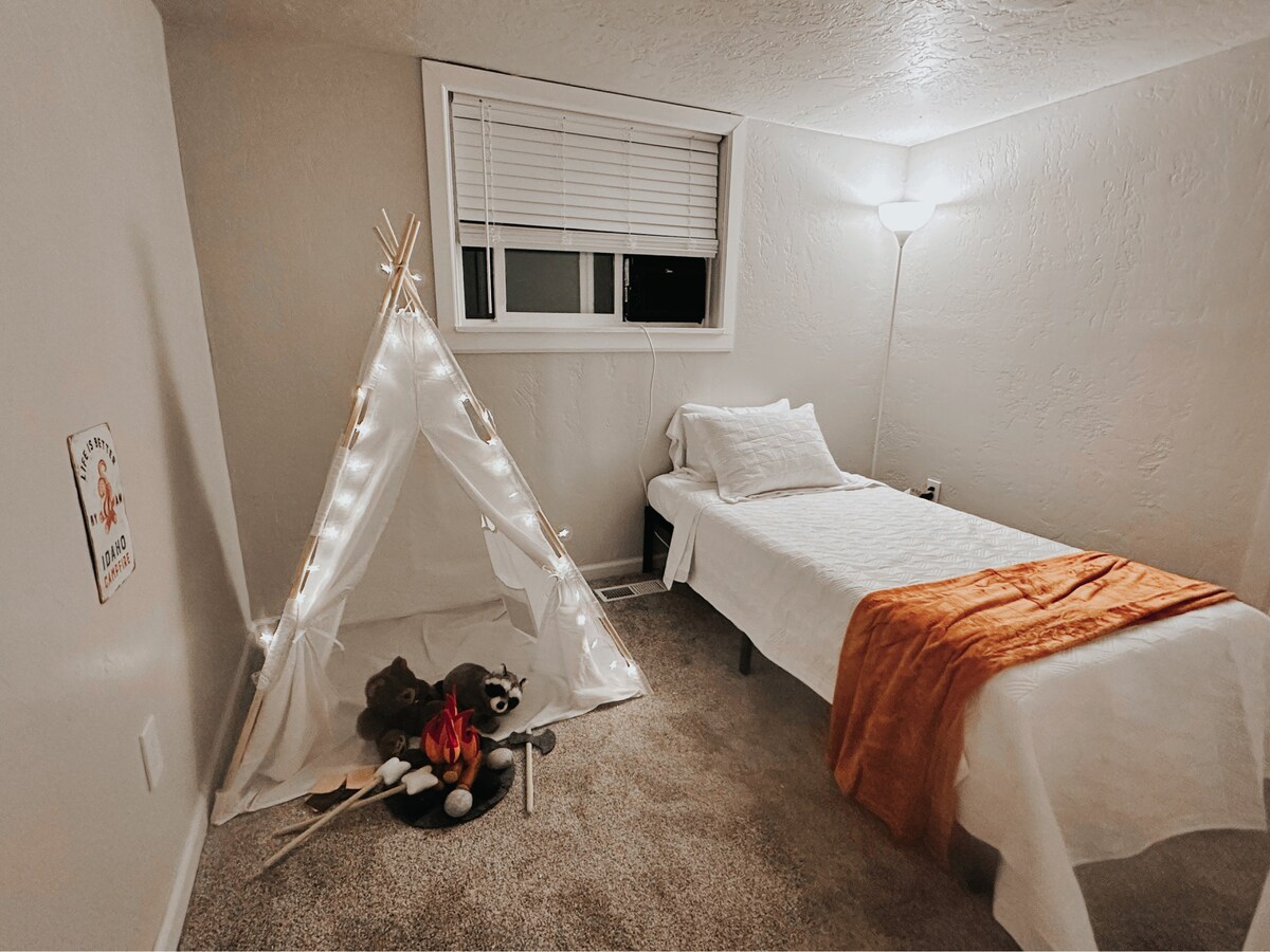 A cozy kids' play area features a lighted tent adorned with string lights and plush toys arranged to create a playful atmosphere. A single bed with a white coverlet is positioned nearby, illuminated by a standing lamp, while a window provides natural light.