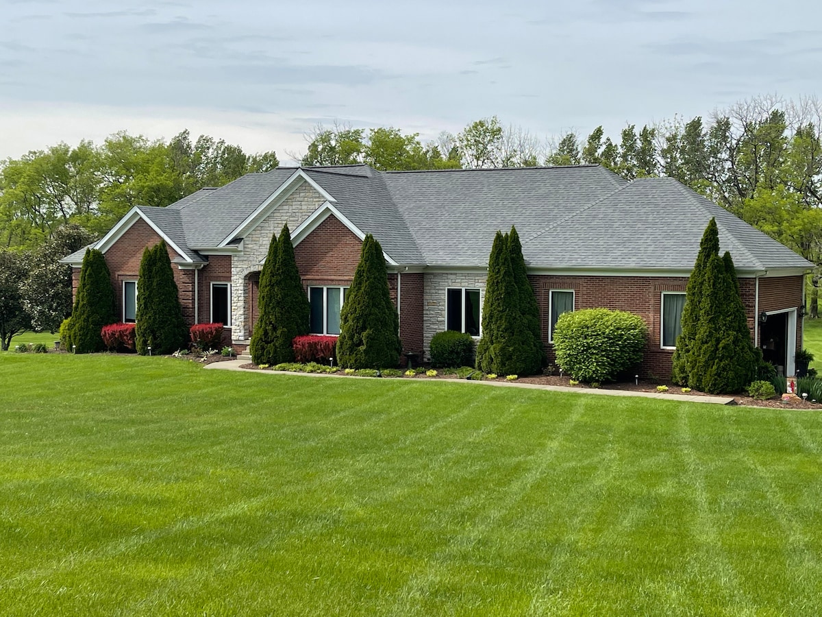 A spacious brick house is surrounded by lush green lawns, featuring neatly trimmed hedges and vibrant flowering plants. Large windows are present throughout the structure, allowing natural light to brighten the interior.