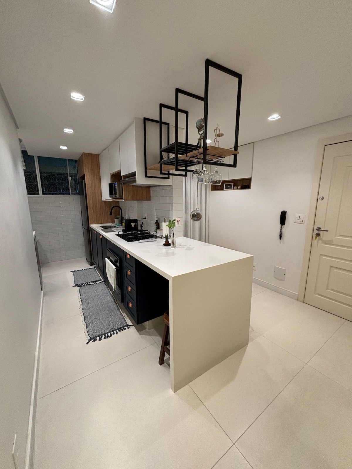A modern kitchen is visible, featuring sleek cabinetry with a combination of wood and dark finishes. A central island includes seating and a gas stove. Bright lighting fixtures hang above, while a window allows natural light to enter, illuminating the clean, minimalist design.