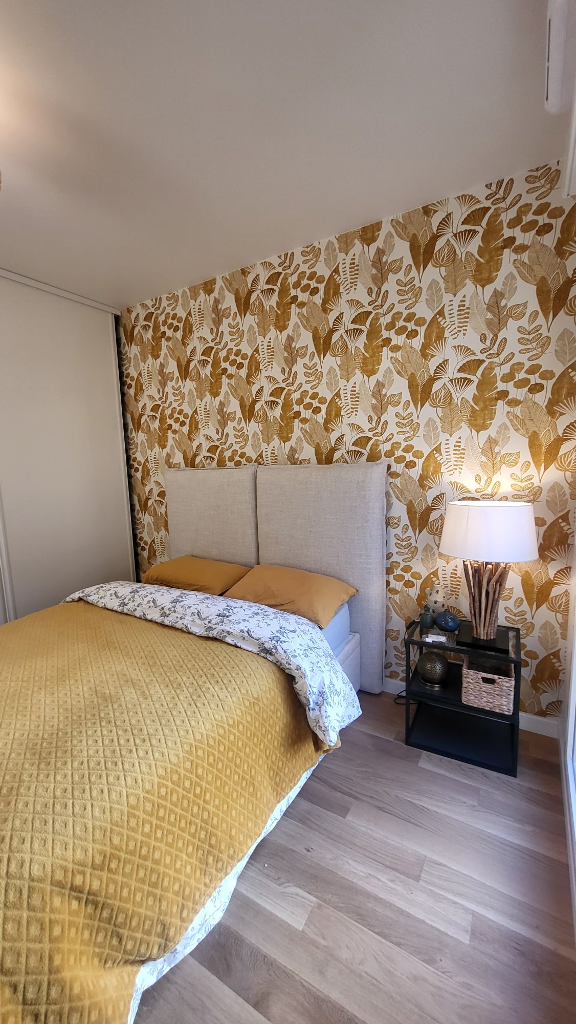 A cozy bedroom features a light-colored headboard against a botanical-patterned wallpaper. A yellow quilted throw covers the spacious bed, while a bedside table with a lamp adds warmth. Natural light softly fills the room, enhancing the calm atmosphere.