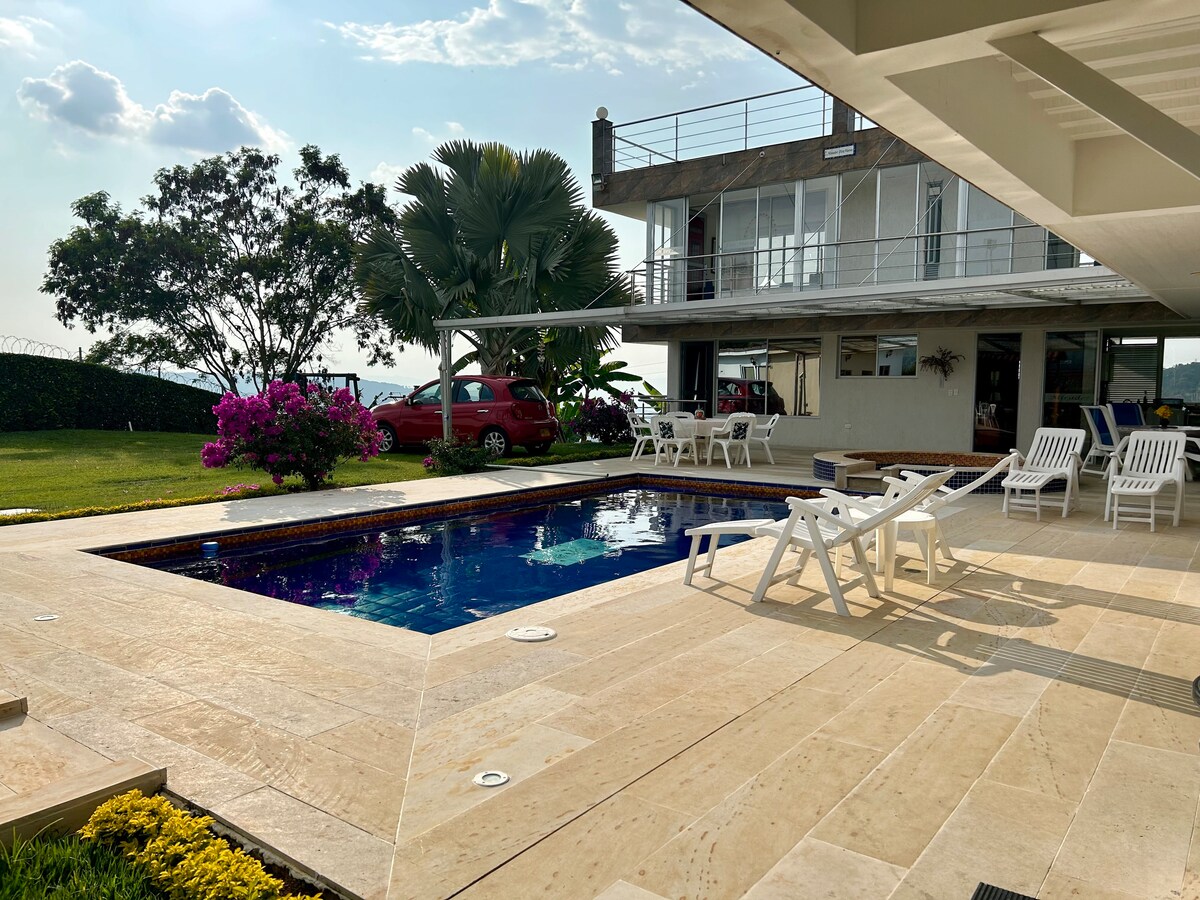 A outdoor area features a rectangular pool surrounded by natural stone tiles. White lounge chairs are positioned beside the water, and colorful flowers and greenery enhance the serene environment. A large two-story building is visible in the background, offering views of the surrounding landscape.