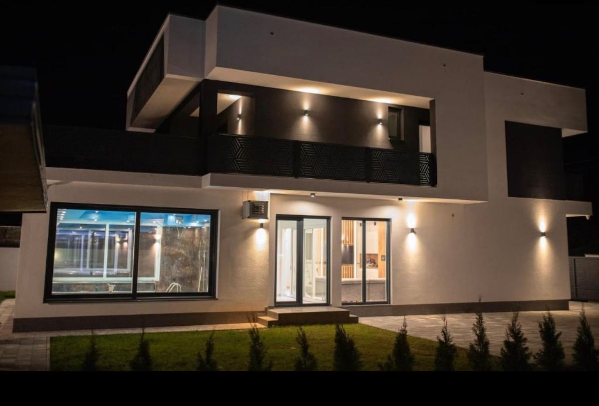 The modern villa exterior is illuminated at night, highlighting its clean lines and architectural design. Large glass windows reflect the surrounding features, while the balcony above offers a glimpse of outdoor seating. Soft lighting enhances the inviting atmosphere of the property.