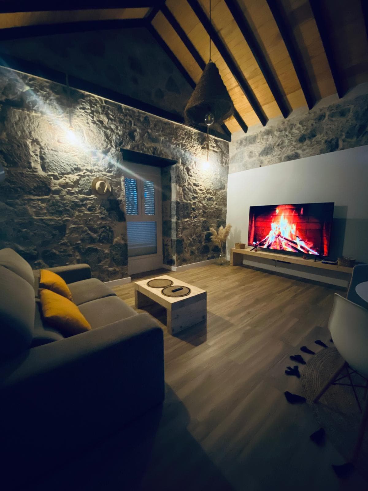 A cozy living area features a stone wall and a wooden ceiling, softly illuminated by wall lights. A modern television displays a fireplace image, while a comfortable sofa is contrasted by orange cushions. A minimalist coffee table completes the inviting space.