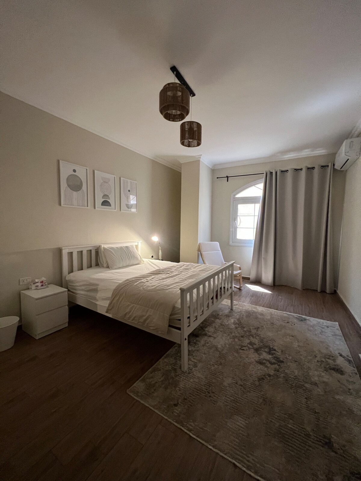 A well-lit bedroom features a spacious bed with neutral bedding, paired with a bedside table. Artwork adorns the wall above the bed, and a cozy armchair is positioned near a window dressed with light curtains. A soft rug adds comfort to the wooden floor.