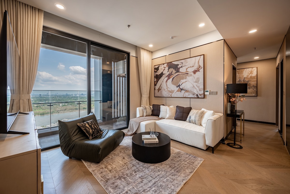 The living room showcases large glass doors that open to a scenic view of the Saigon River. A spacious white sofa and a unique dark lounge chair are arranged around a round coffee table atop a soft area rug. Artwork frames the room, adding a contemporary touch.