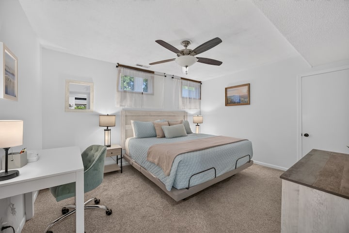 A king bed with end tables, dresser, closet, tv, ceiling fan, and a dedicated workspace. There is a bathroom next door. The windows have room darkening shades and light filtering curtains.