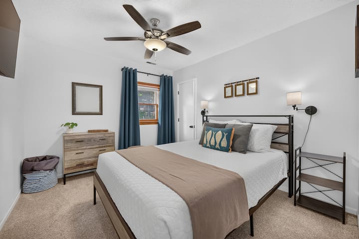 A queen bed with closet, dresser, tv, end table, ceiling fan, and a bathroom next door. The window has light filtering mini blinds and room darkening curtains.