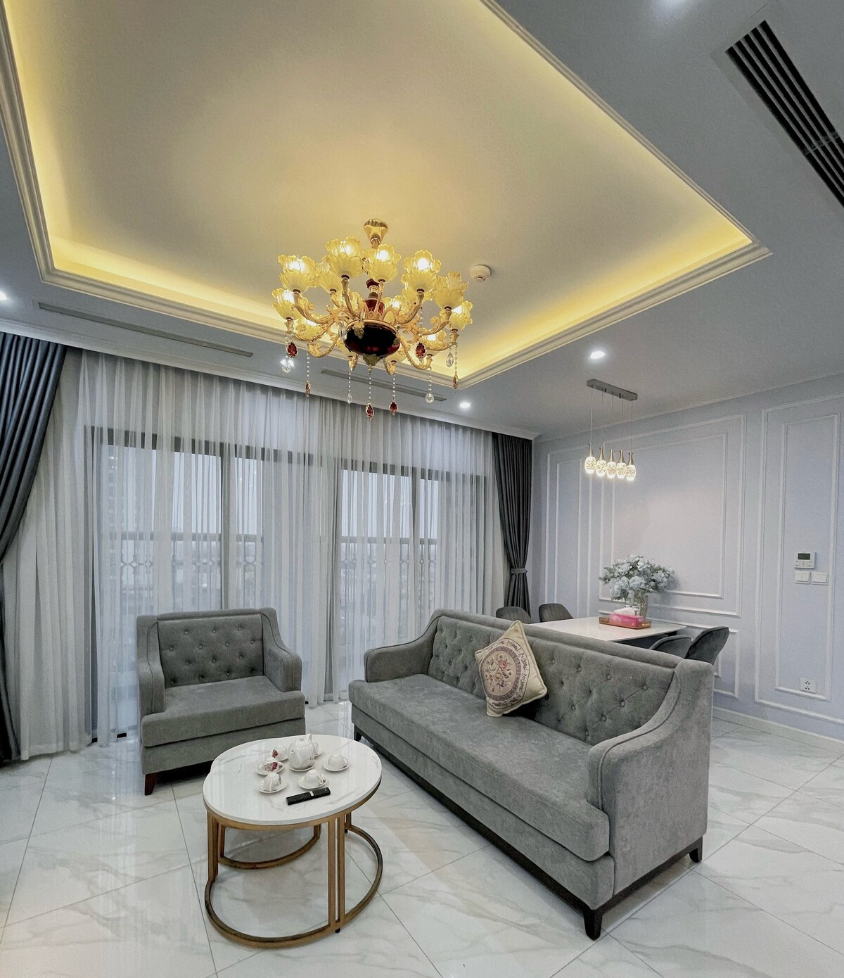 A stylish living area features a plush gray sofa and an accent chair, complemented by a round coffee table. Large windows are draped with light curtains, allowing natural light to brighten the space. A chandelier hangs from the ceiling, adding an elegant touch.
