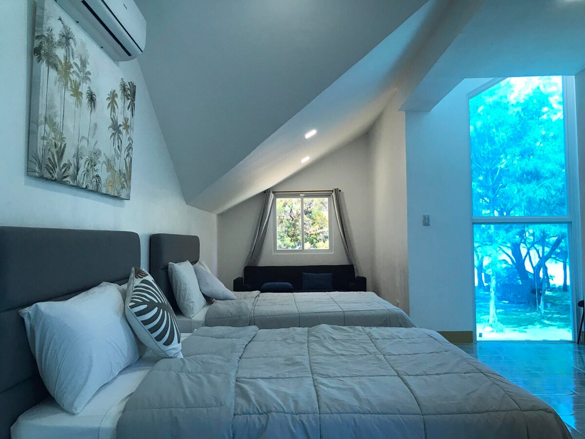 A bright sleeping area features two spacious beds with neutral-colored bedding. Large windows allow natural light to enter, framing a view of greenery outside. Light-colored walls and a tropical wall painting contribute to a calming atmosphere.