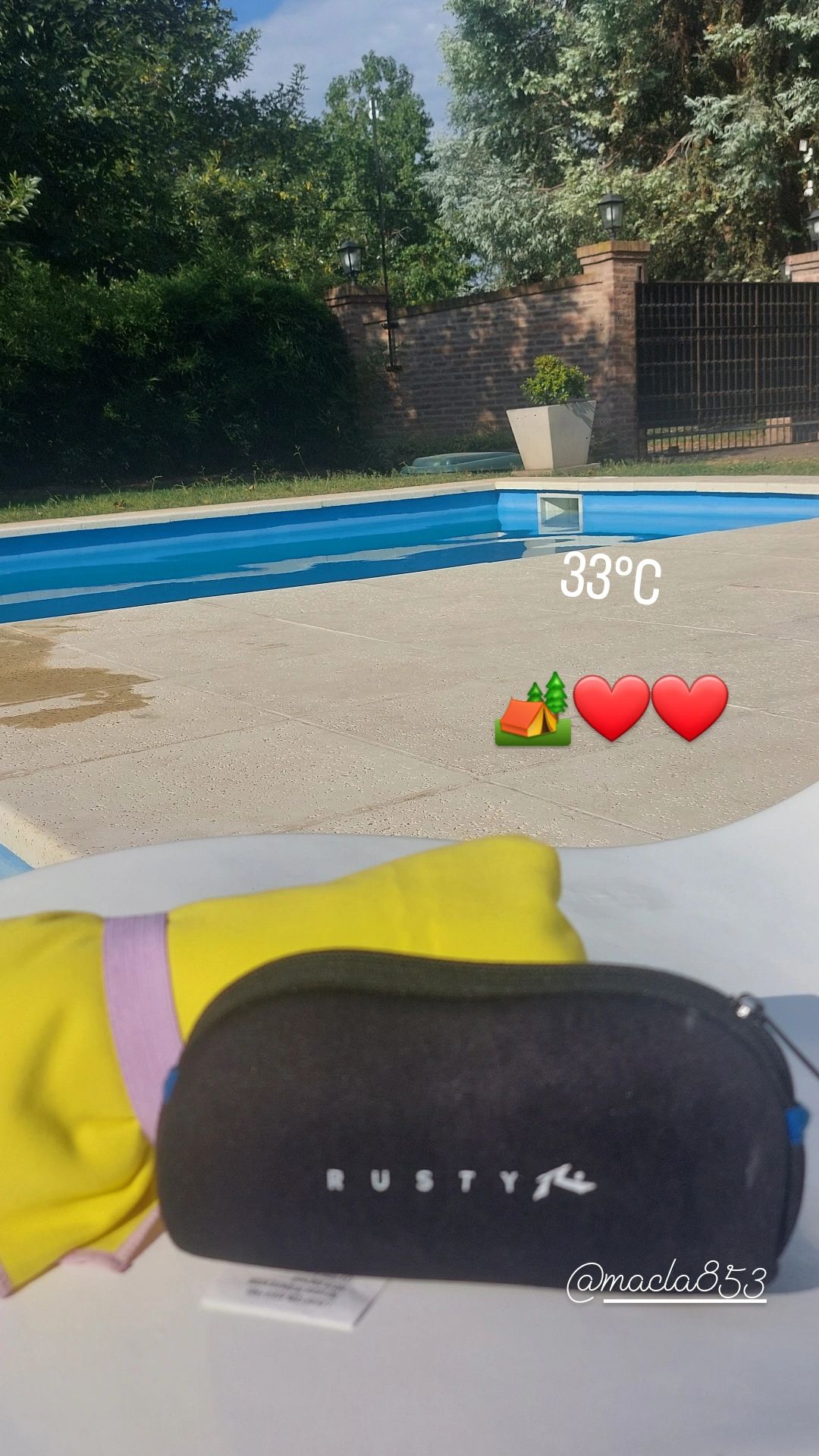 A serene swimming pool is shown, with clear water reflecting blue skies. A colorful towel and a black case are placed on a nearby surface, suggesting a relaxing day by the poolside. Lush greenery surrounds the area, adding to the tranquil ambiance.