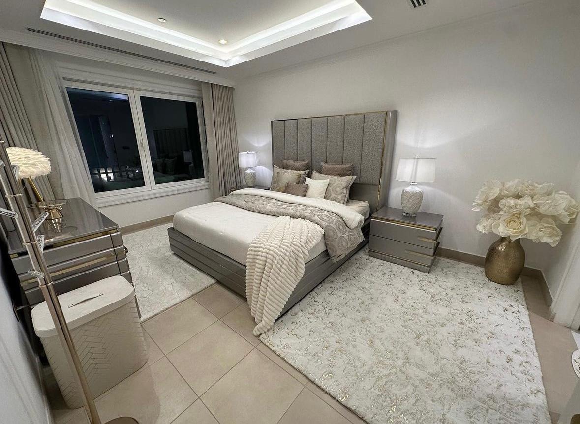 The bedroom features a spacious bed with elegant linens and decorative pillows, flanked by bedside lamps. A large window allows natural light to fill the room, which is adorned with a soft area rug and stylish furniture, including a mirrored dresser and an accent chair.