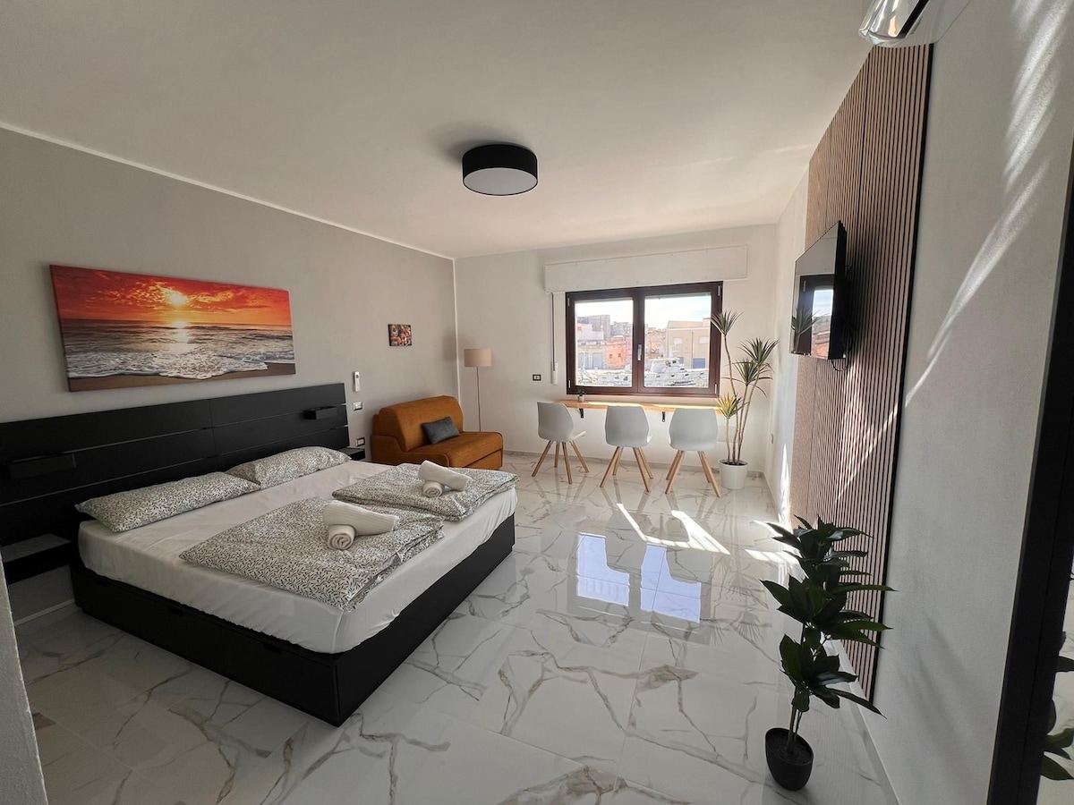 A spacious room is highlighted by large windows allowing ample natural light. It contains a large bed with neatly arranged linens, a comfortable seating area with a sofa, and a dining space with chairs. A decorative plant adds a touch of greenery to the modern design.