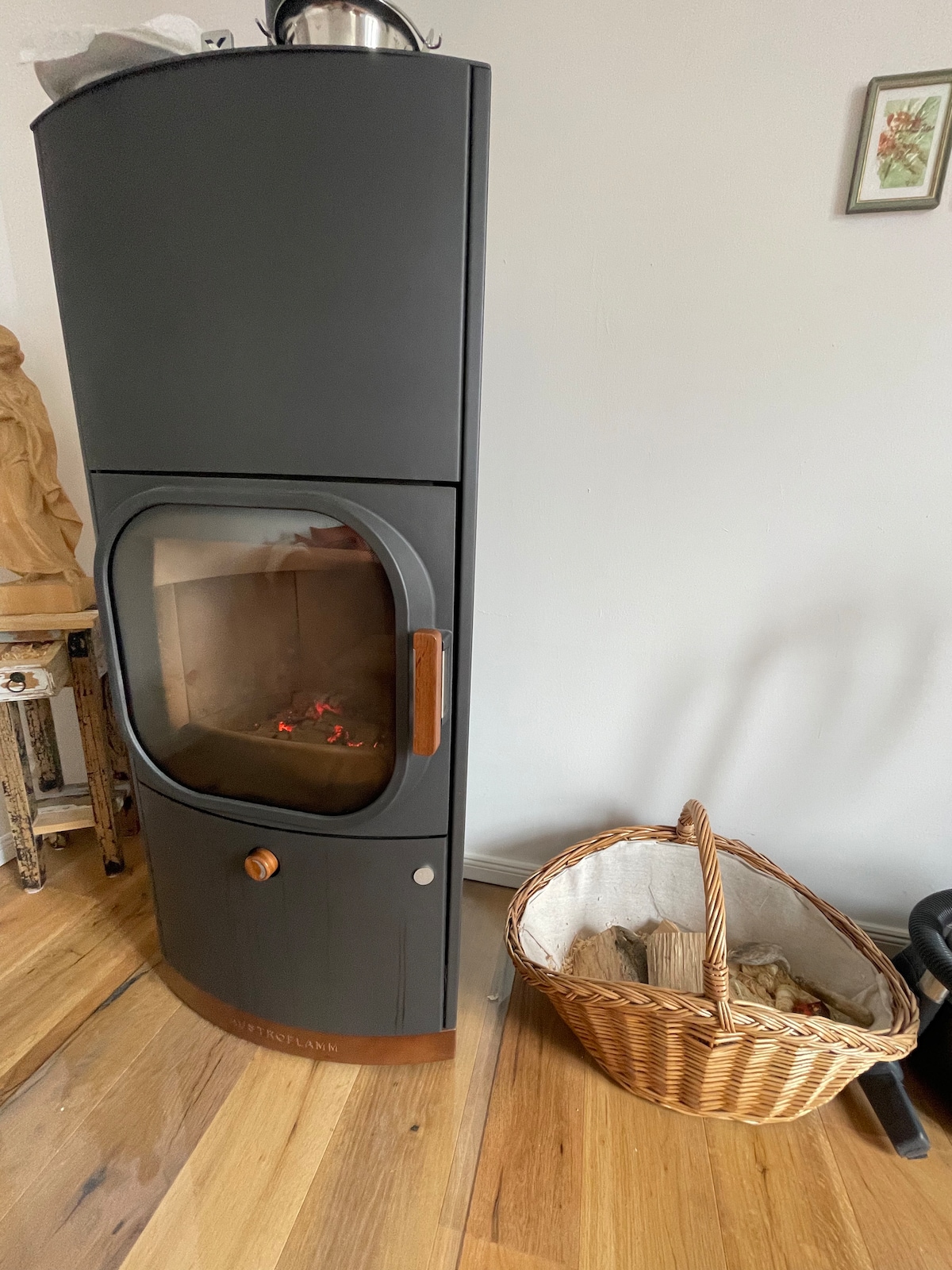 A modern wood-burning stove is positioned against a light-colored wall, featuring a glass door that reveals flickering flames inside. A woven basket filled with firewood is placed nearby on a hardwood floor, adding a functional touch to the cozy space.