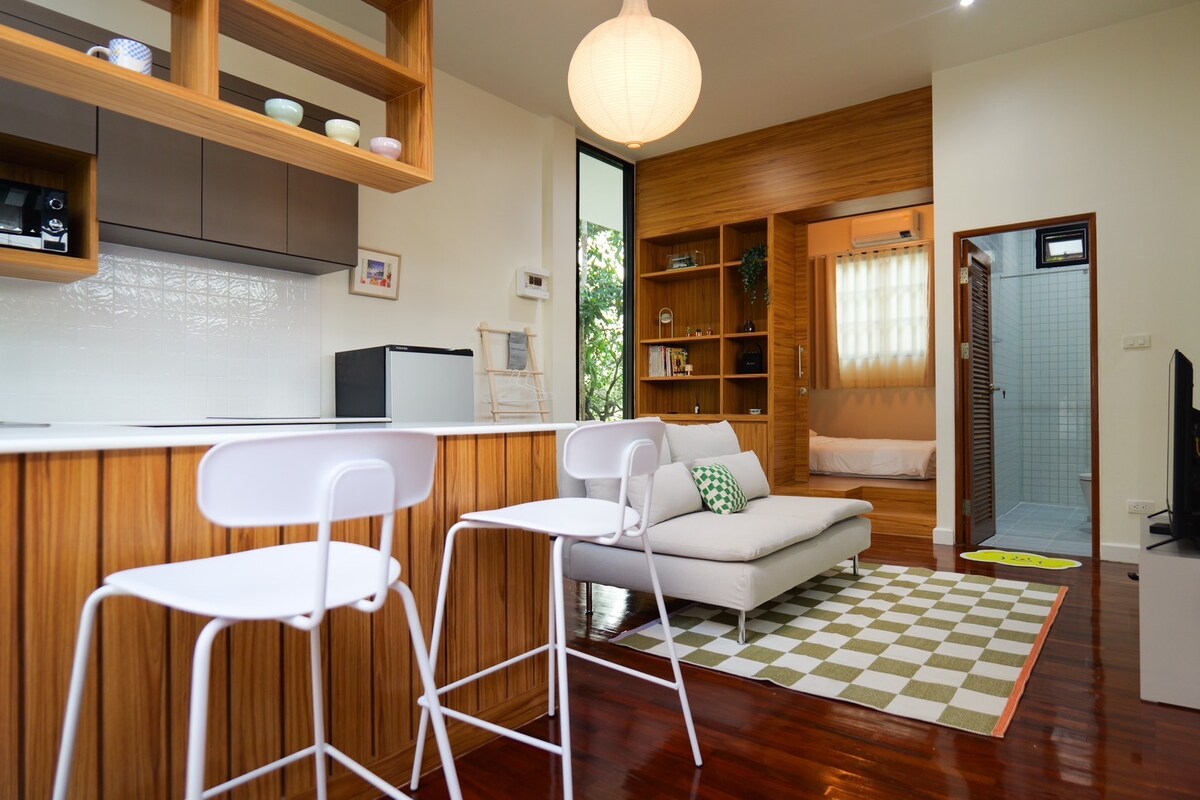 A cozy living area features a comfortable gray sofa placed on a patterned rug. A small wooden shelf showcases decorative items, while a kitchenette with modern appliances is situated nearby. Natural light filters through large windows, illuminating the warm wooden elements of the space.