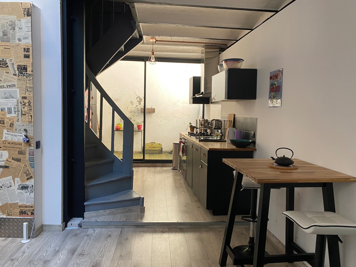A modern kitchen is visible, featuring sleek black cabinetry and stainless steel appliances. A wooden table with two white chairs is positioned nearby. A staircase leads to the upper levels, while natural light filters in through a window at the back of the space.
