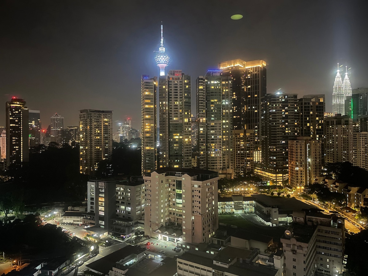 A panoramic view of Kuala Lumpur at night is displayed, featuring illuminated skyscrapers and distinctive landmarks. The iconic Petronas Twin Towers and KL Tower are visible, surrounded by city lights that reflect the vibrant urban environment.