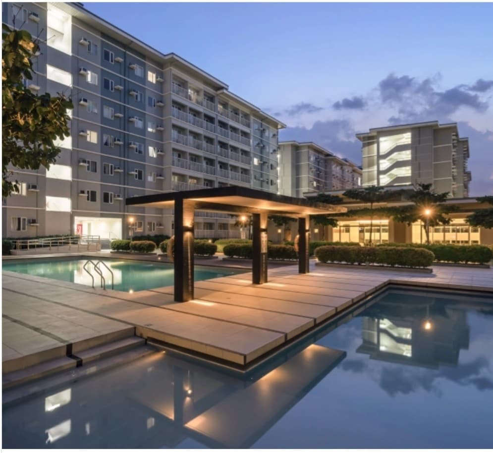 A modern swimming pool is framed by a well-lit wooden cabana, with steps leading into the water. Surrounding trees and neatly trimmed bushes enhance the area. Multi-story buildings provide a contemporary backdrop against a serene twilight sky.