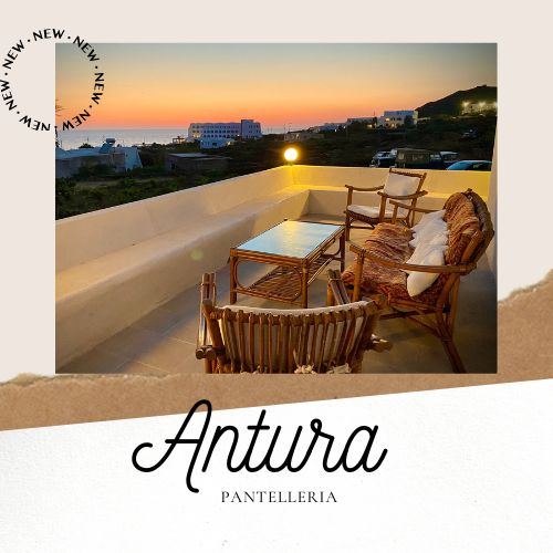 An outdoor terrace is furnished with a table and two wicker chairs, providing a comfortable space for relaxation. A soft glow from a nearby light enhances the welcoming setting. The view includes a serene sunset over the sea, creating a peaceful atmosphere.