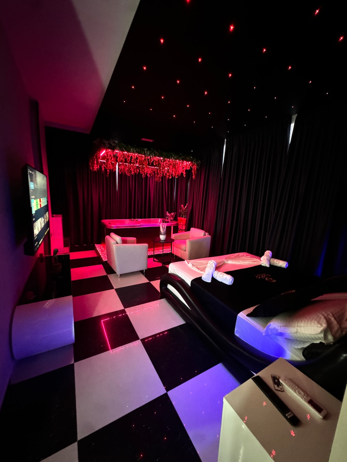 A modern room features checkered black and white flooring and is illuminated by soft red lights. A spacious bed is complemented by a chic seating area with two armchairs. A crib with vibrant decorative accents is nearby, and a large television is mounted on the wall.