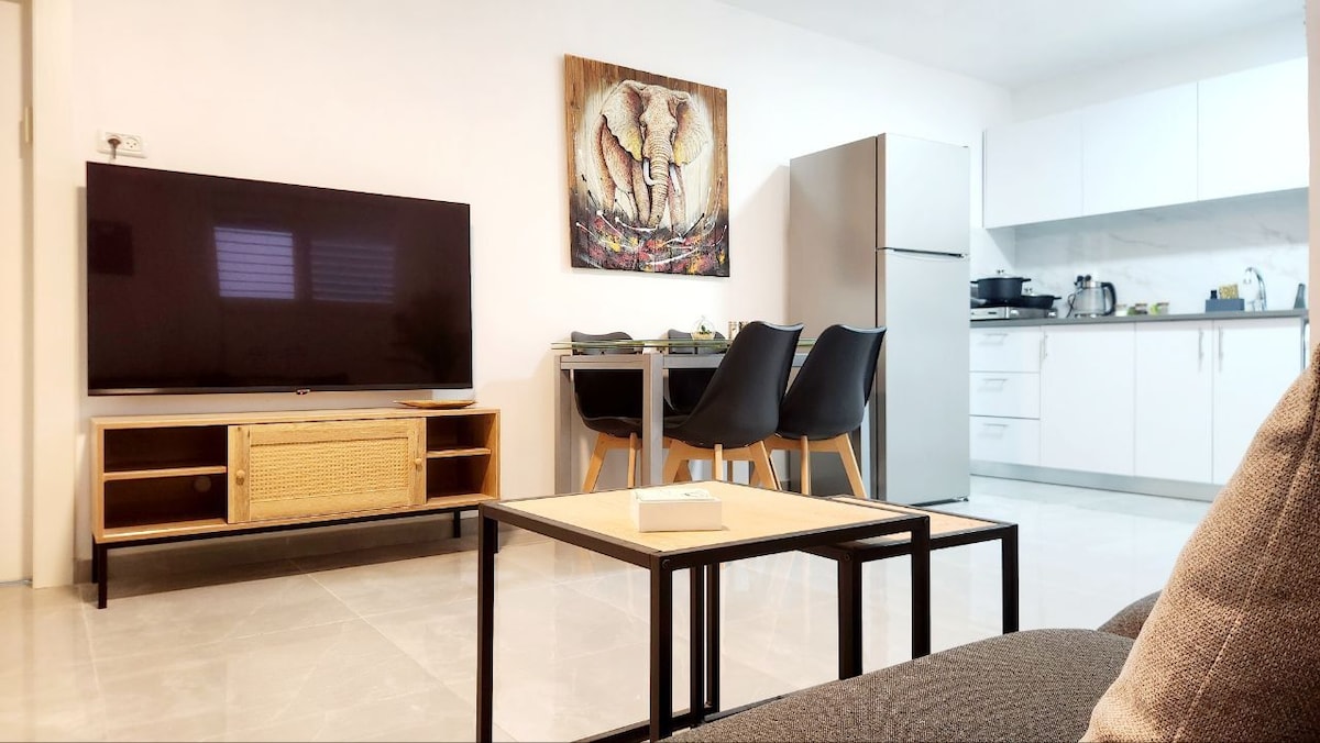 A modern living area is shown, featuring a wall-mounted television above a low coffee table. A stylish dining table with four black chairs is visible alongside a fully equipped kitchen. Light-colored cabinetry complements the space, while an artwork of elephants adorns the wall.