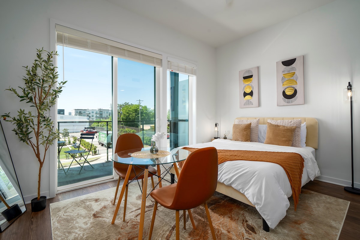 A bright bedroom features a comfortable queen bed adorned with decorative pillows and a textured throw. A small glass dining table is set with two chairs, complemented by a large window offering natural light and outdoor views, creating an open and airy ambiance.