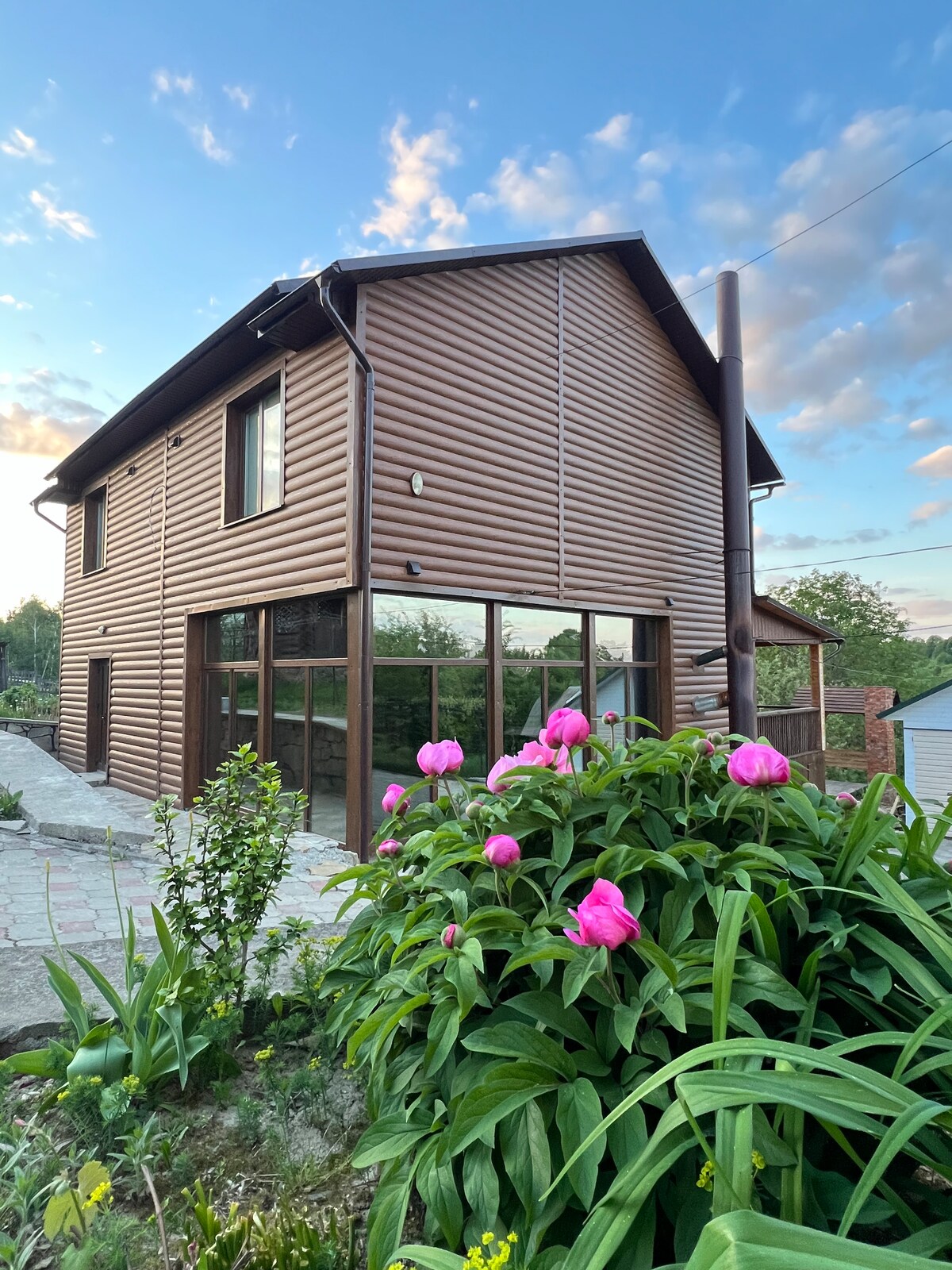 The exterior of a two-story wooden house is captured, featuring large glass doors that blur the line between indoors and outdoors. Vibrant pink flowers bloom in the foreground, while a pathway leads to the entrance, framed by green foliage and a clear sky.
