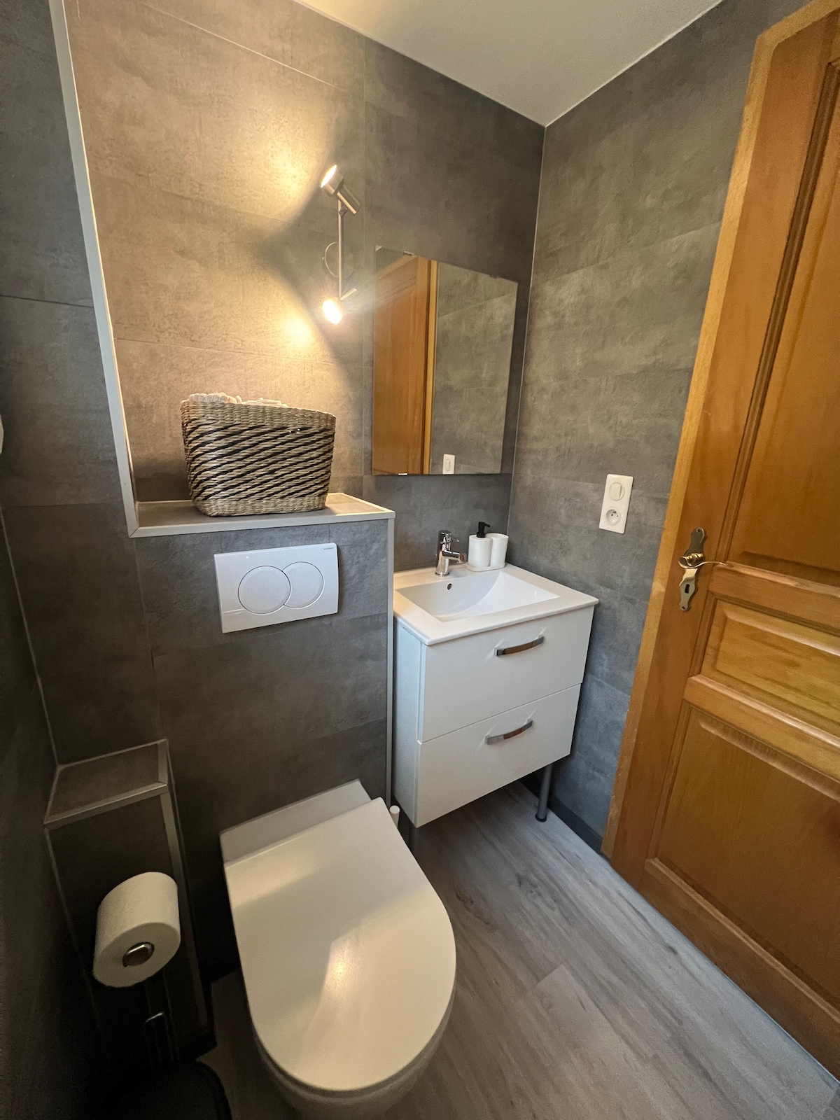 The bathroom is designed with contemporary grey tiling. A modern toilet and a compact washbasin with storage are included. A mirror reflects the warm light from a wall fixture. A basket sits atop the cupboard, and essential toiletries are neatly arranged.