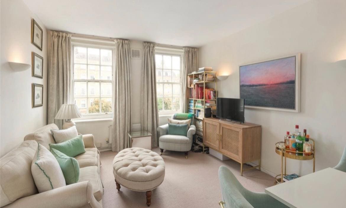 A bright living room features large windows with sheer curtains allowing natural light. A comfortable sofa and an armchair are accompanied by a tufted ottoman. A TV sits on a mid-century style cabinet, while a small dining table adds functional space.