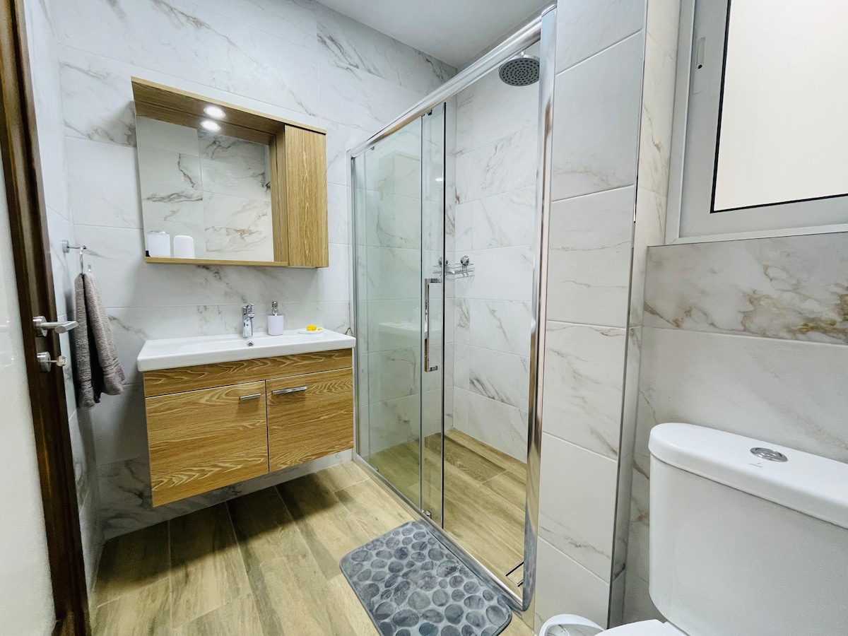 A modern bathroom features a glass shower stall and a wooden vanity with a large mirror above. Light-colored marble walls complement the neutral flooring. Bath essentials are neatly arranged, and a soft mat lies in front of the shower for added comfort.