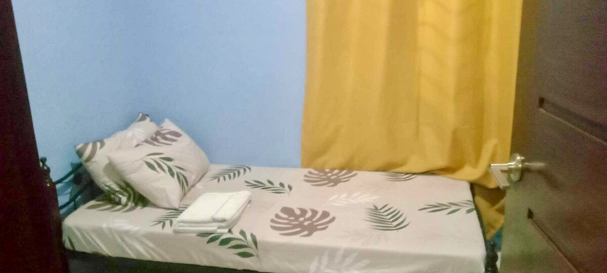 Eenpersoonsbed met plafondventilator en kleine kast. 