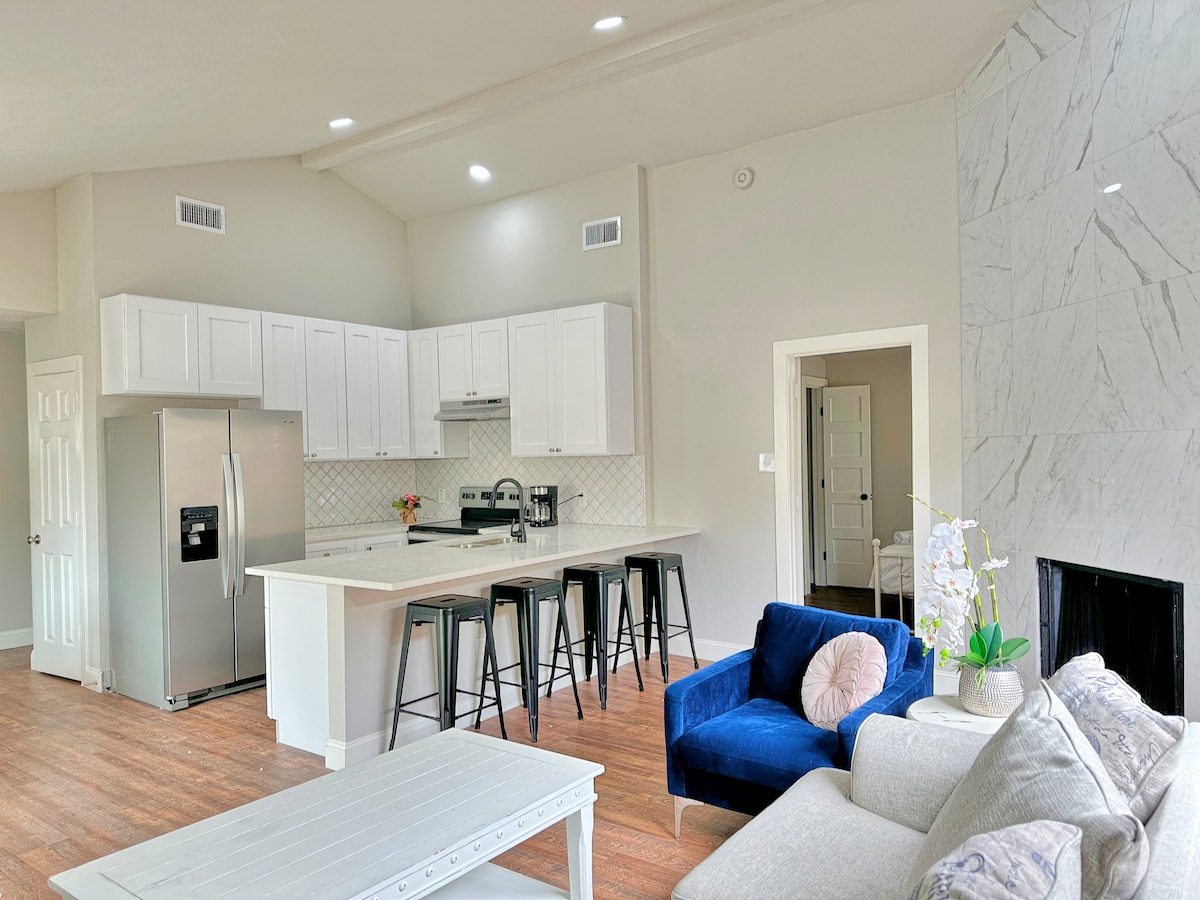 A modern kitchen showcases white cabinetry and stainless steel appliances, with a bar area featuring tall black stools. The open living space includes a blue couch and a light gray sofa, complemented by natural light from the large windows.