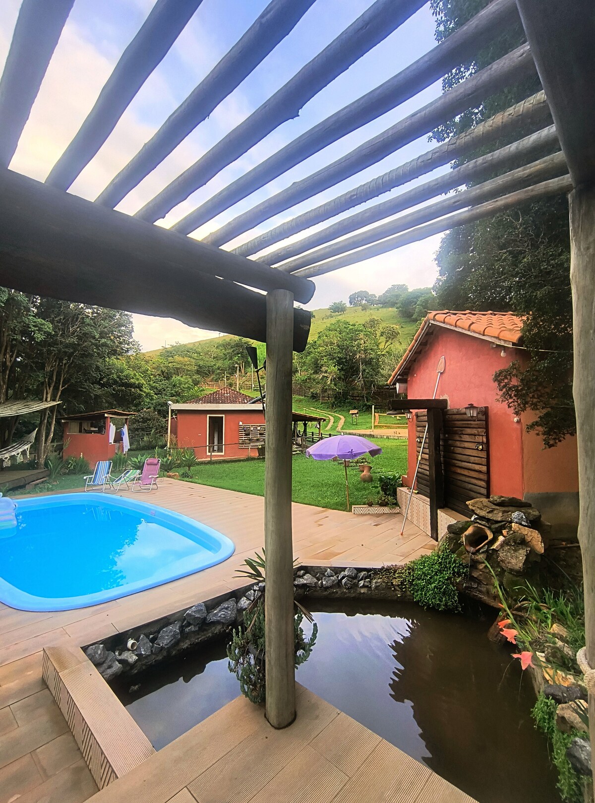 A serene outdoor area features a pool surrounded by a wooden deck. Lush green hills rise in the background, with colorful lounge chairs arranged on the lawn. A small pond with rocks adds to the natural ambiance, enhancing the overall peaceful setting.
