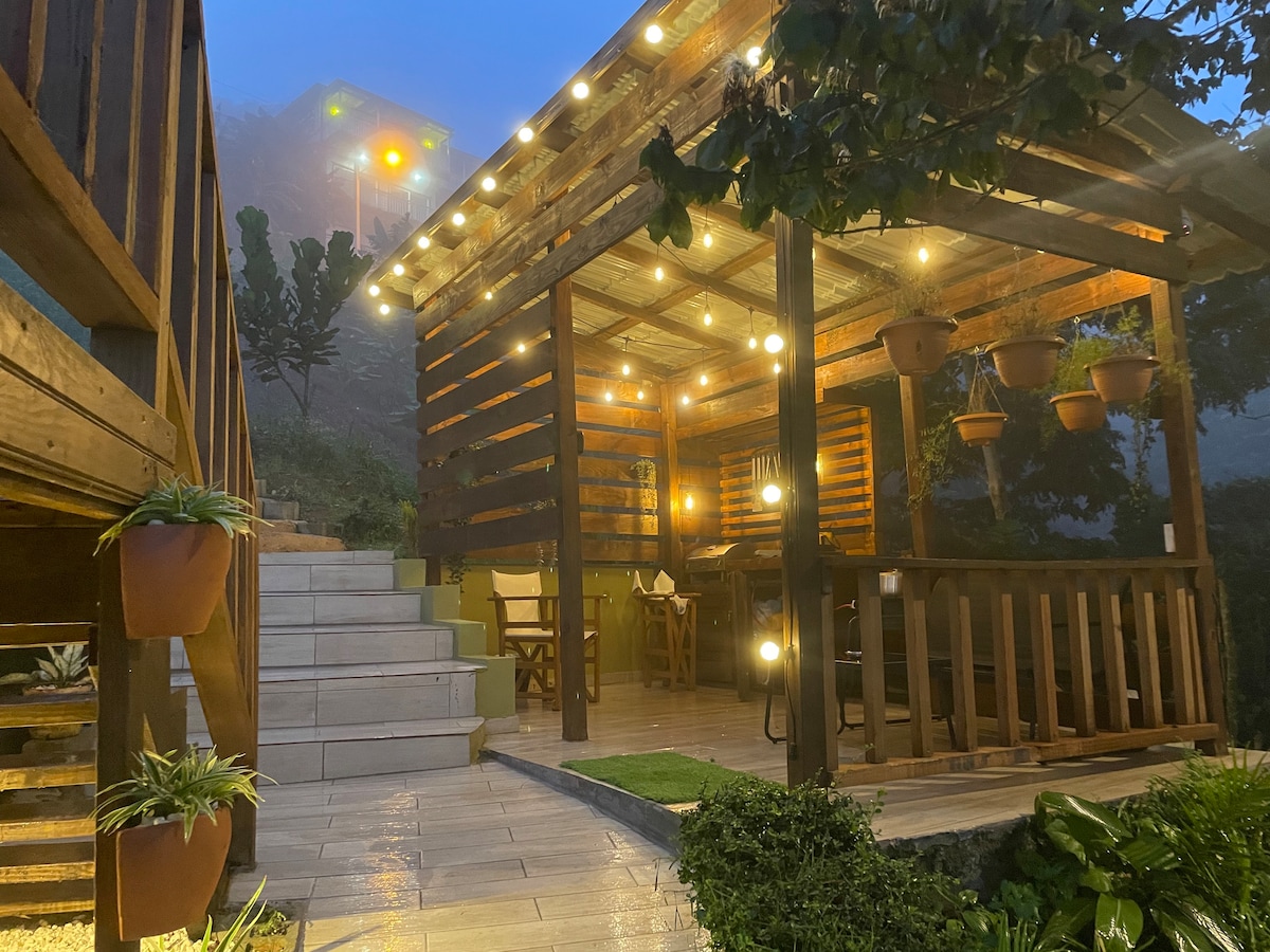 A charming deck area is illuminated with warm lights, showcasing wooden beams and hanging plants. Steps lead up from a tiled path, bordered by lush greenery, towards a cozy dining space under shelter that invites relaxation amidst the surrounding mountain mist.