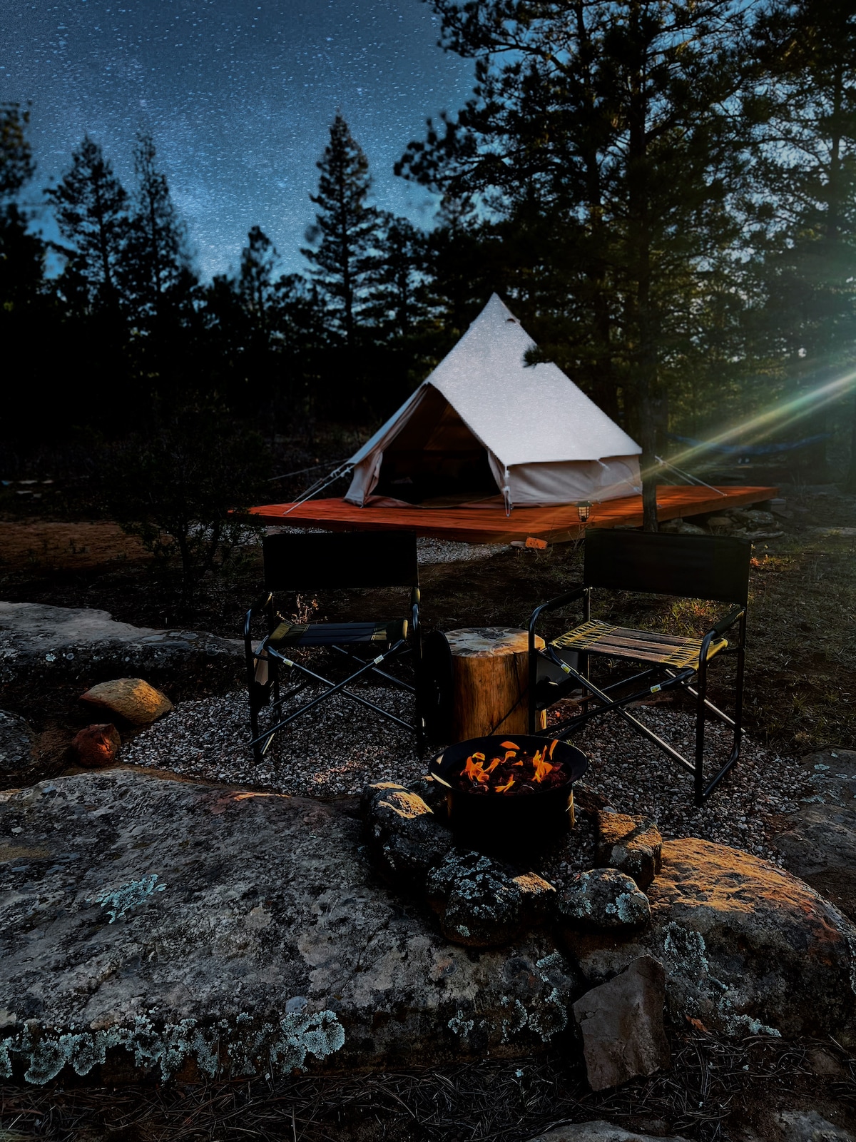 A bell tent is illuminated under a starry sky, situated on a rocky landscape. Nearby, a fire pit with flames is surrounded by two black chairs, offering a cozy outdoor seating area framed by trees.