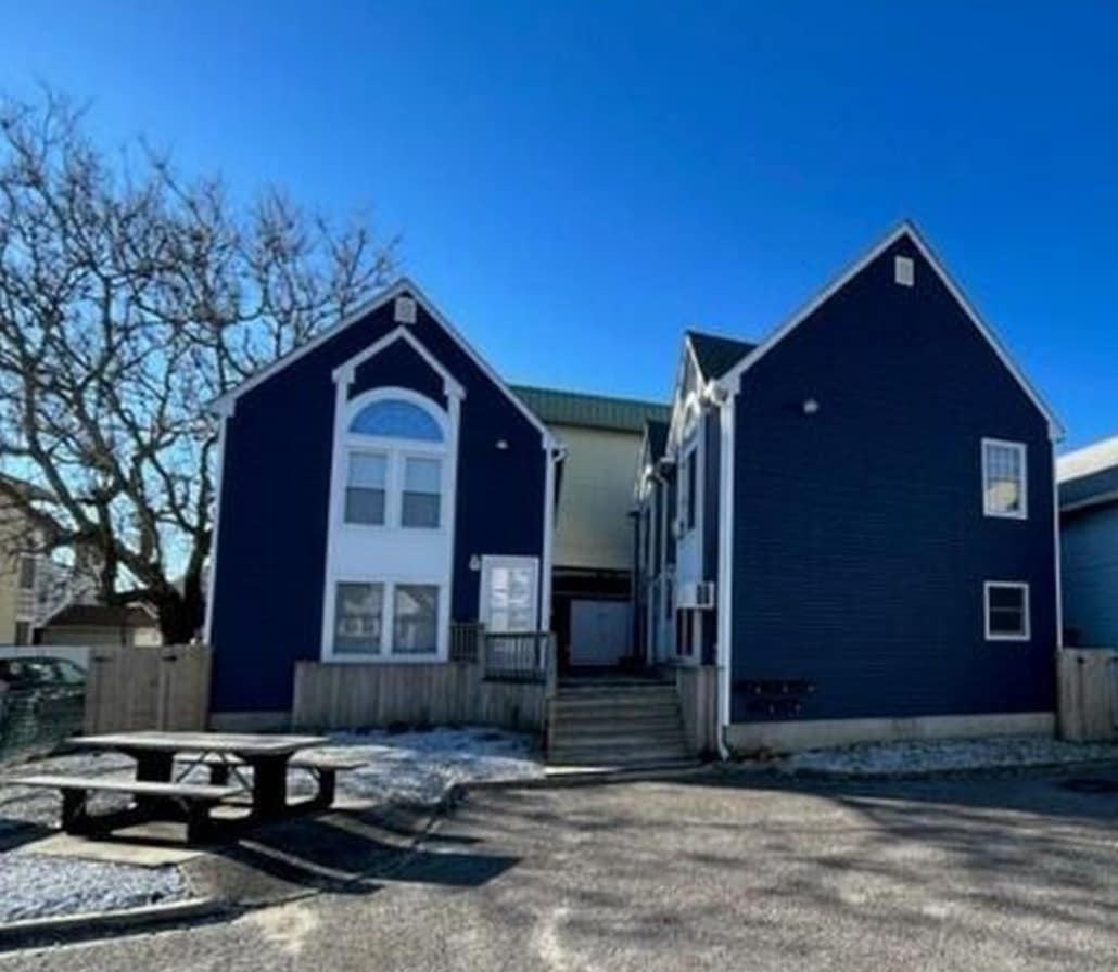The exterior of the two-story condo is shown, featuring dark blue siding and large windows. A picnic table is positioned in the foreground. The entrance is marked by a small set of stairs leading up to the main door.