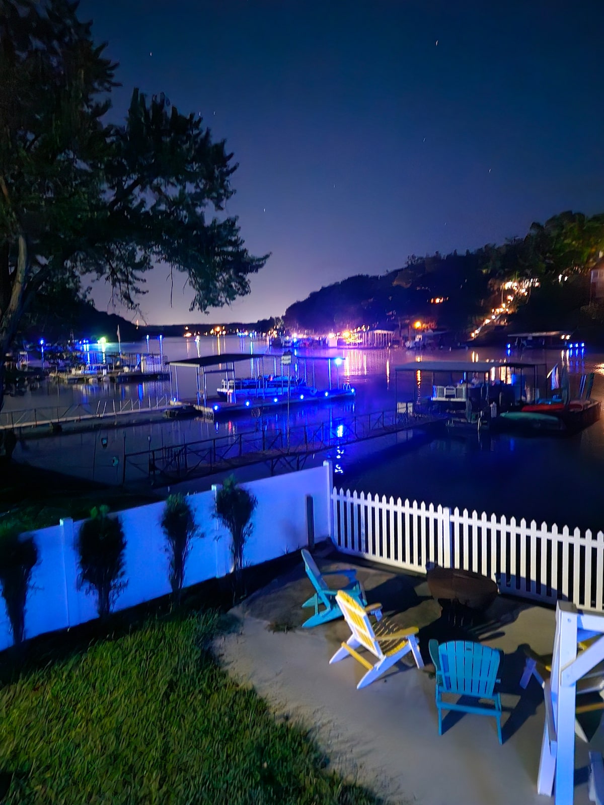 The image captures a lakeside scene at night, featuring illuminated docks with blue lighting reflecting on the water. A wooden fence lines the foreground, where Adirondack chairs are positioned, creating a welcoming spot for relaxation amidst the tranquil setting.