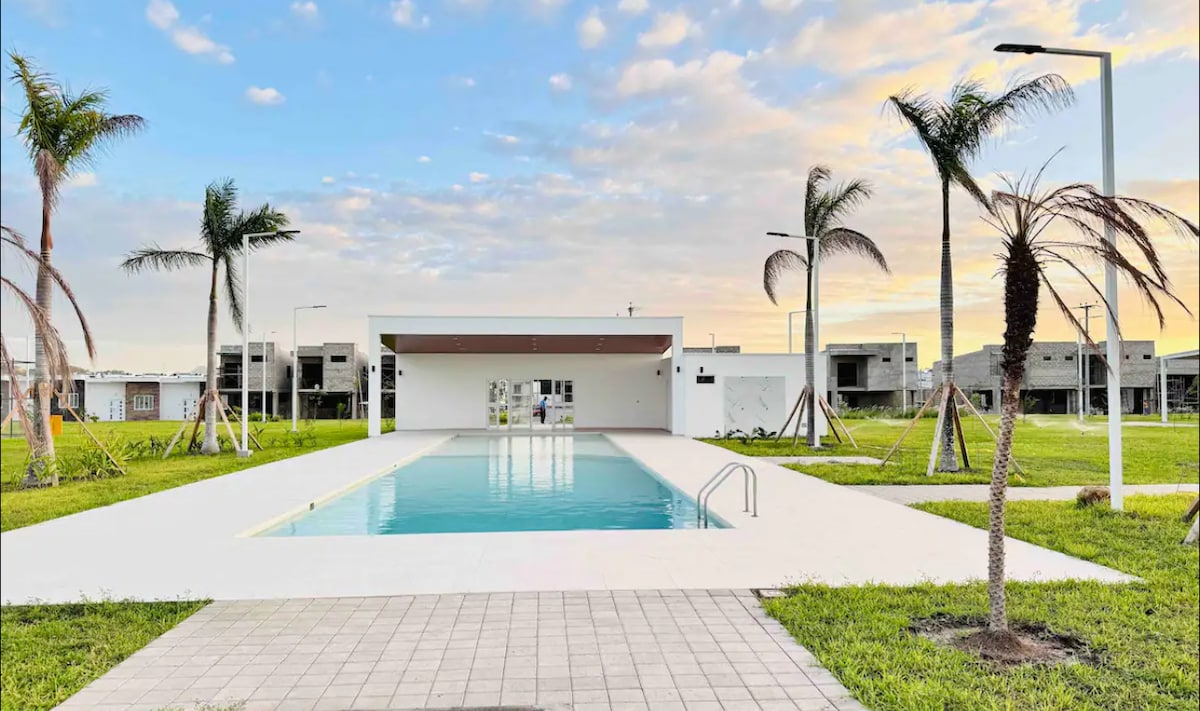 A serene pool area is surrounded by neatly trimmed grass and palm trees. The pool is framed by a spacious deck, reflecting the soft colors of the sky during sunset. A building in the background complements the modern aesthetic.