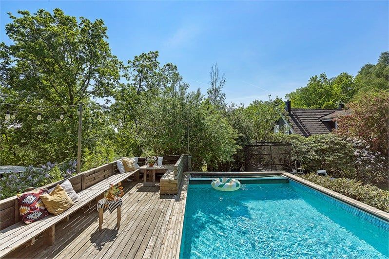 An outdoor patio features wooden seating arranged around a central table, surrounded by lush greenery. A rectangular swimming pool is situated nearby, with a floatation device resting on the water. Sunlight illuminates the scene, enhancing the relaxing atmosphere.