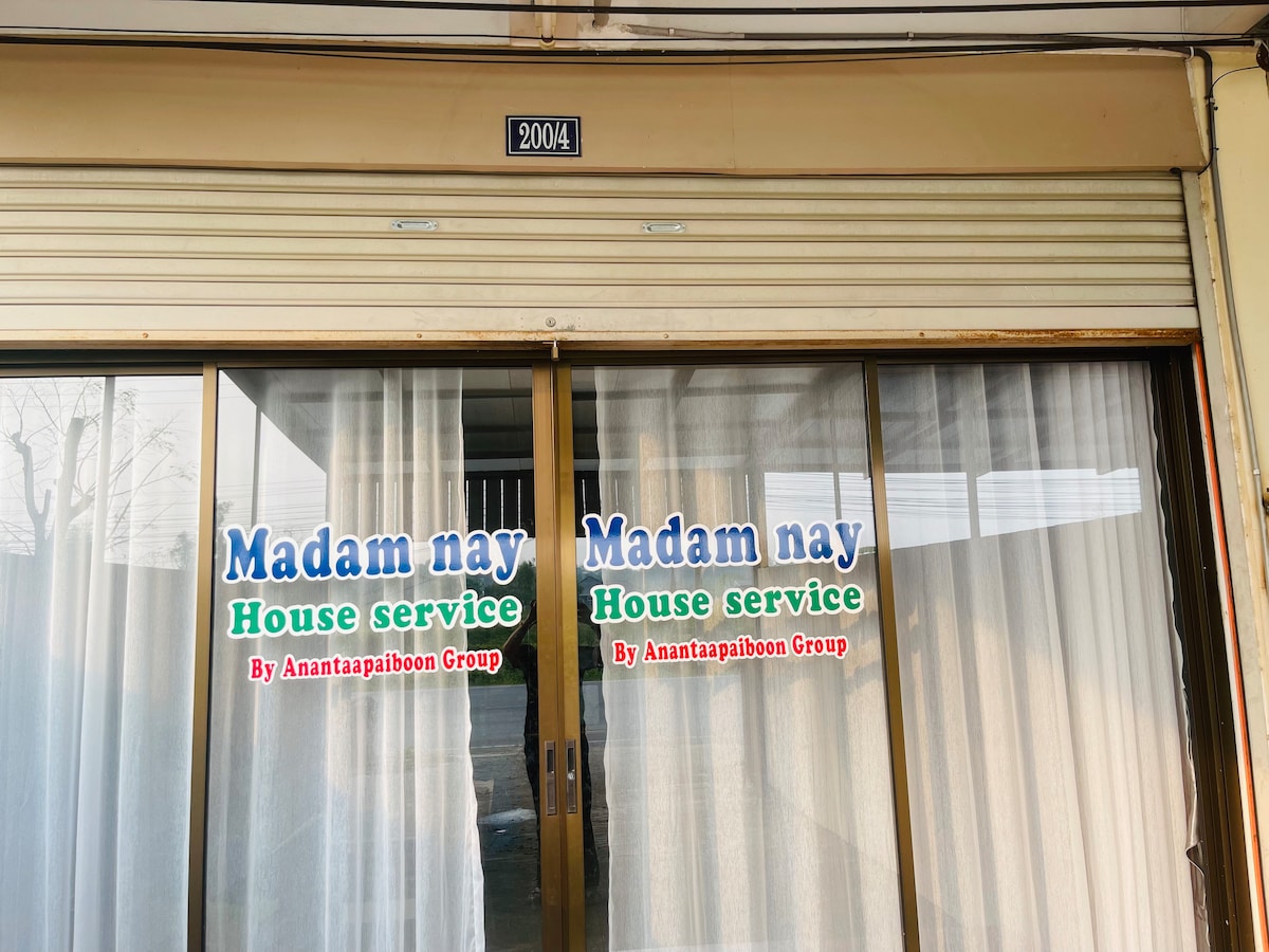 A shopfront is showcased with large glass doors marked with 'Madam nay House service' signage, indicating the establishment's name. White curtains are seen behind the glass, allowing soft light to filter through.