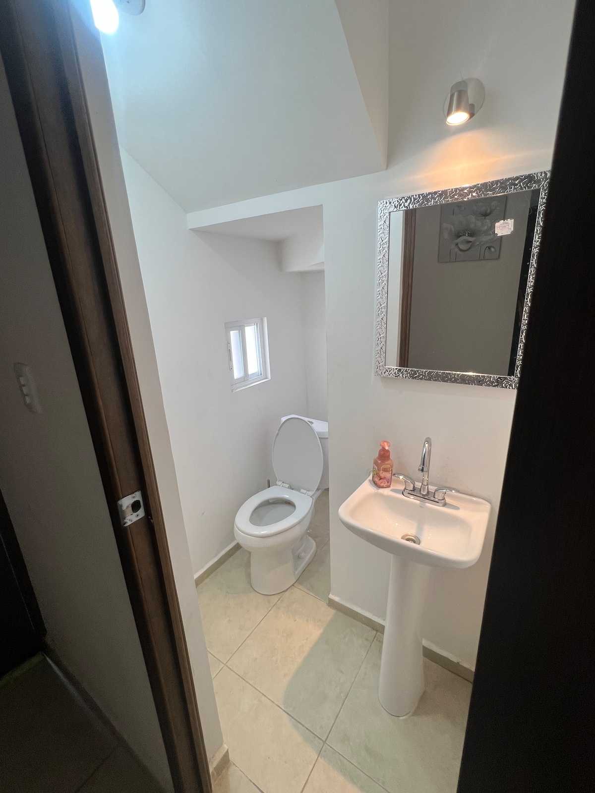 A compact bathroom is featured with a sleek sink positioned beside a modern toilet. Natural light enters through a small window, and a decorative mirror adds a touch of elegance. Neutral tiles extend across the floor and walls, enhancing a clean environment.
