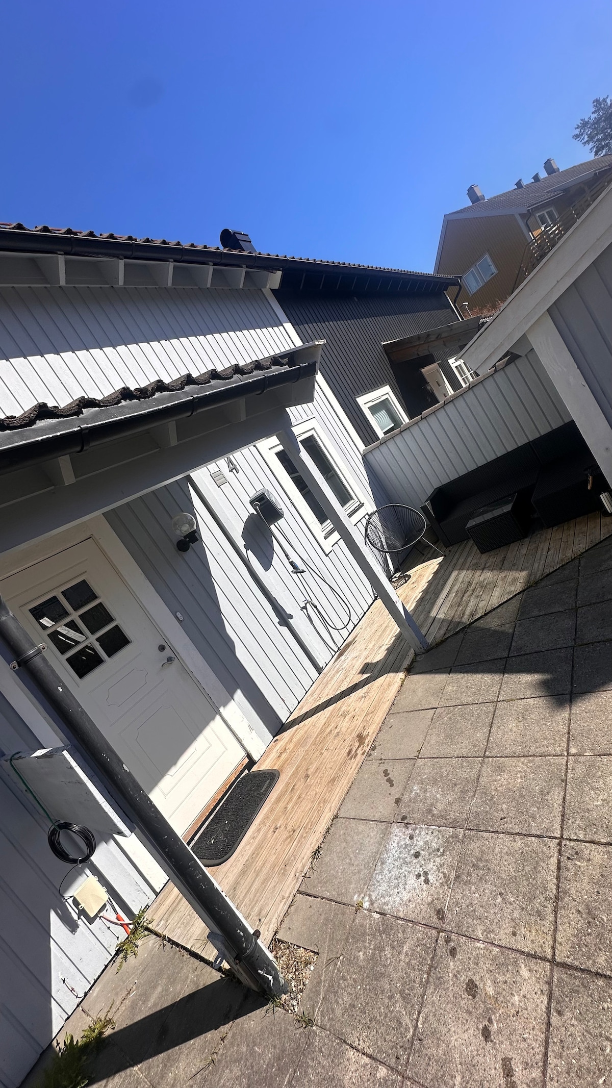 An outdoor area is presented with a patio featuring wooden decking and paved tiles. A white entrance door is visible, alongside a modern seating arrangement. Clear blue skies provide ample natural light, enhancing the inviting atmosphere of the space.