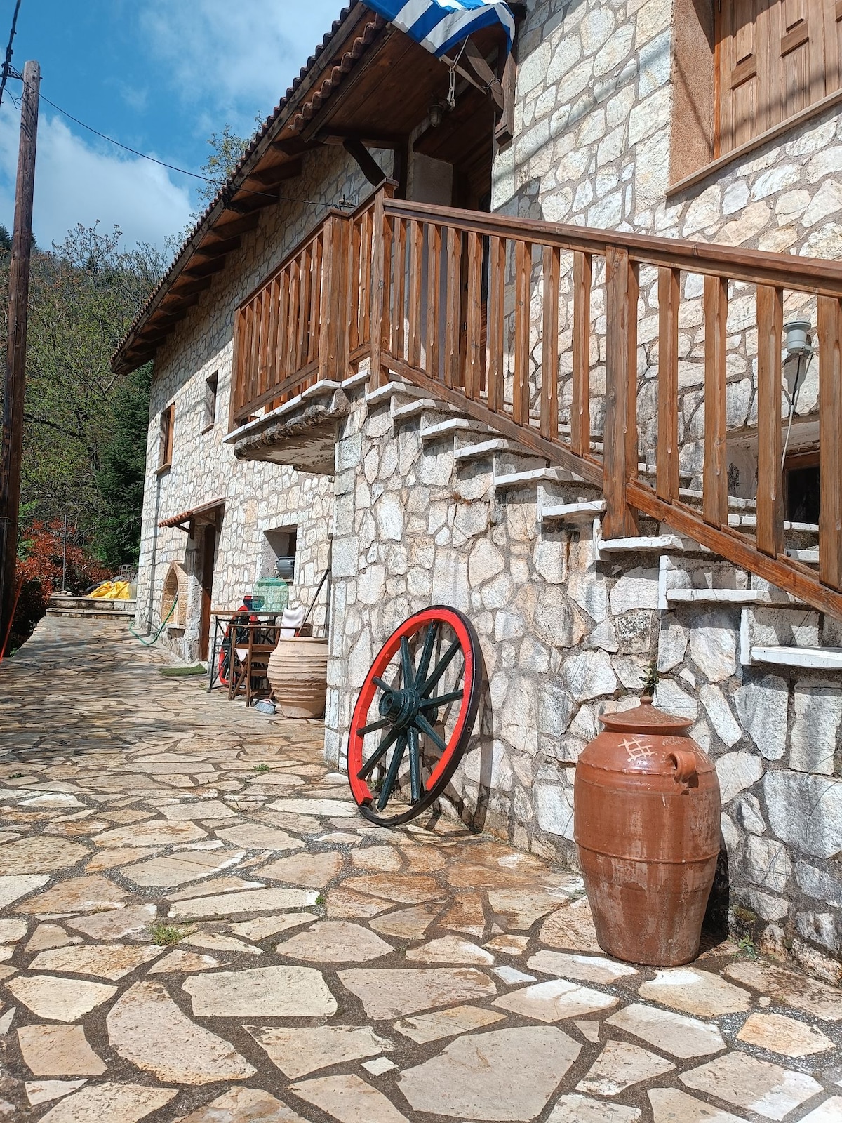 A stone exterior features a rustic design complemented by wooden balconies and steps. A cobblestone pathway is lined with terracotta pots and a large wooden wheel, inviting exploration of the outdoor space surrounded by greenery.