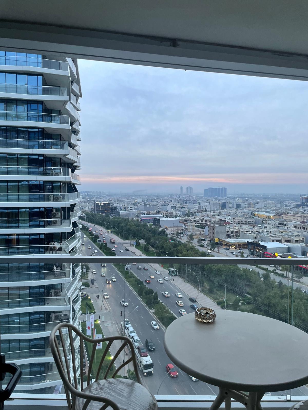 A modern balcony view overlooks a bustling cityscape, featuring roadways filled with cars and buildings in the distance. Glass paneling reflects the light, while a small round table with a decorative item sits near a pair of chairs, inviting relaxation.