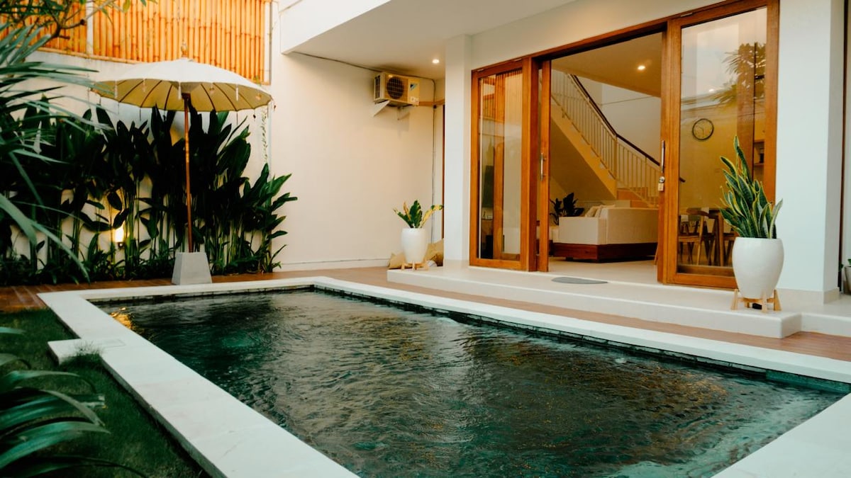 A private swimming pool is framed by lush greenery and modern decor. Bright wooden doors open to reveal a spacious living area, while a shaded umbrella stands beside the pool, adding a touch of comfort to the outdoor space.