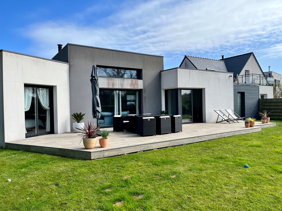 The exterior of the modern house features a spacious wooden deck with outdoor seating. Potted plants are arranged around the area, and two lounge chairs are positioned on the grass. Large windows and a patio door provide views into the bright interior. A clear blue sky is visible above.