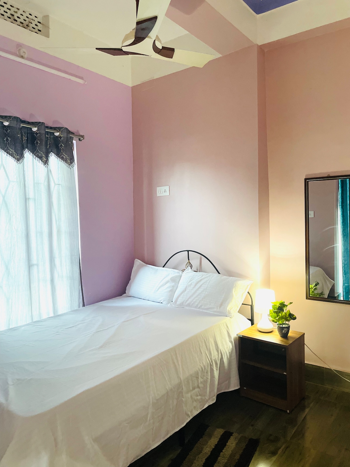 A bright room features a spacious double bed with crisp white bedding against a soft pink wall. Natural light filters through a large window with sheer curtains. A small bedside table holds a plant and a lamp, while a mirror adds depth to the space.